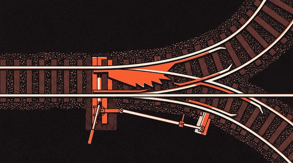A railway switch seen from above, one track dividing into two across dark gravel, its moving blade caught mid-throw.